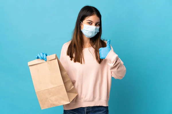 マスクでコロナウイルスから保護し 何か良いことが起こっているので 親指で青の背景に隔離された食料品の買い物袋を保持若い女性 — ストック写真