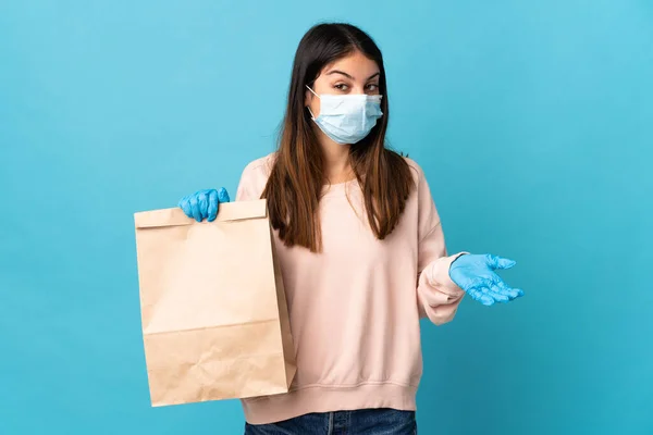 マスクでコロナウイルスから保護し 青い背景に隔離された食料品の買い物袋を保持している若い女性は肩を持ち上げながら疑問ジェスチャーを作ります — ストック写真