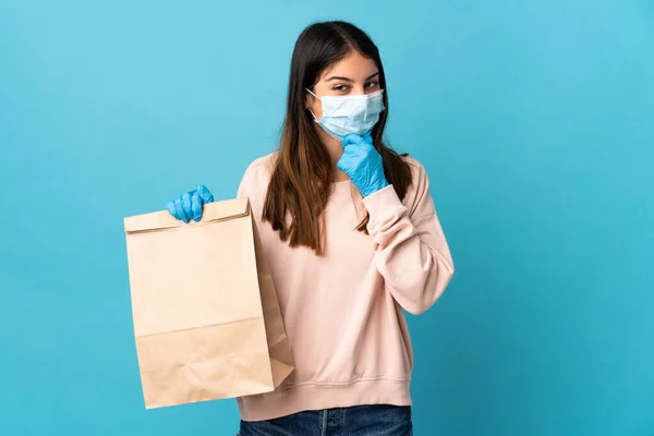マスクでコロナウイルスから保護し 青の背景に隔離された食料品の買い物袋を保持する若い女性考えと見て側 — ストック写真