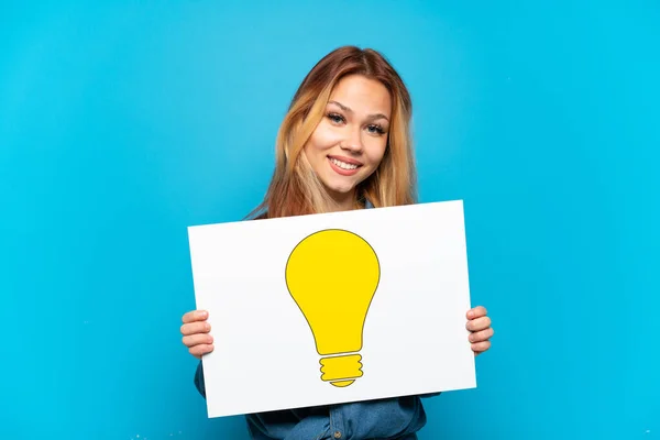 Teenager Girl Isolated Blue Background Holding Placard Bulb Icon Happy — Zdjęcie stockowe