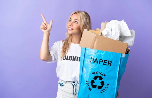 若いロシア人女性を保持リサイクルバッグフル紙でリサイクルするために隔離された紫の背景に指差す指で素晴らしいアイデア — ストック写真