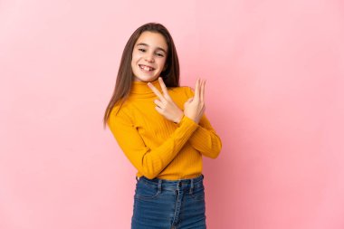 Pembe arka planda izole edilmiş küçük kız gülümsüyor ve zafer işareti gösteriyor