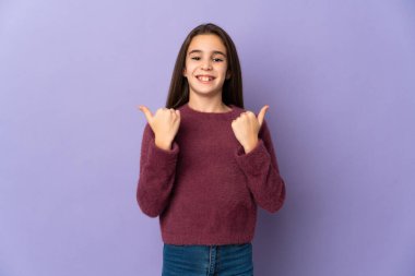 Mor arka planda izole edilmiş küçük bir kız başparmakları havada ve gülümsüyor.