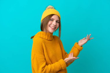 Kışlık ceket giyen genç İngiliz kadın mavi arka planda izole edilmiş. Davet edilmek için ellerini yana uzatıyor.