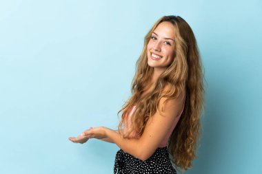 Mavi arka planda izole edilmiş genç sarışın kadın davet etmek için ellerini yana uzatıyor.