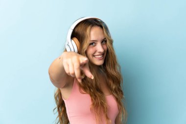 Mavi arka planda izole edilmiş genç sarışın kadın müzik dinliyor.