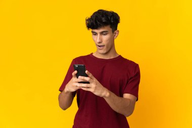 Young Argentinian man isolated on yellow background looking at the camera while using the mobile with surprised expression