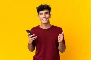 Young Argentinian man isolated on yellow background using mobile phone while doing thumbs up