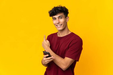 Young Argentinian man isolated on yellow background using mobile phone and pointing back