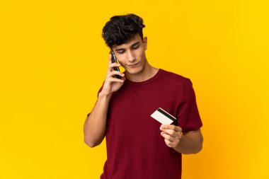 Arjantinli genç adam sarı arka planda izole edilmiş ve cep telefonuyla kredi kartı almış.