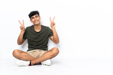 Young Argentinian man sitting on the floor showing victory sign with both hands
