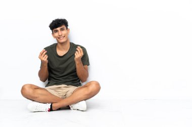 Young Argentinian man sitting on the floor making money gesture