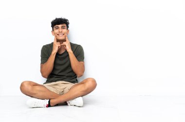 Young Argentinian man sitting on the floor smiling with a happy and pleasant expression