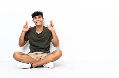 Young Argentinian man sitting on the floor with fingers crossing