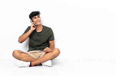 Young Argentinian man sitting on the floor keeping a conversation with the mobile phone with someone