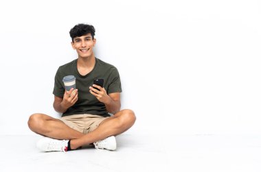Genç Arjantinli bir adam elinde kahve ve cep telefonu ile yerde oturuyor.
