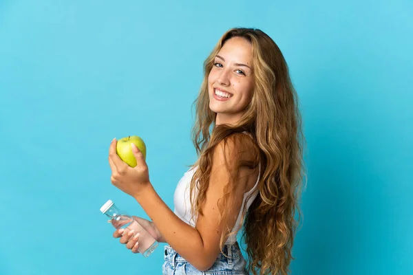 一个金发碧眼的年轻女人 带着一个苹果和一瓶水 被蓝色的背景隔开了 — 图库照片