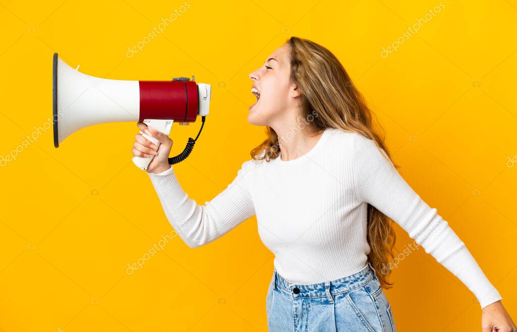 Young blonde woman isolated on yellow background shouting through a megaphone to announce something in lateral position