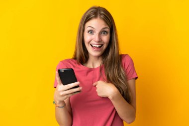 Sarı arka planda cep telefonu kullanan genç bir kadın. Şaşırtıcı bir yüz ifadesi var.