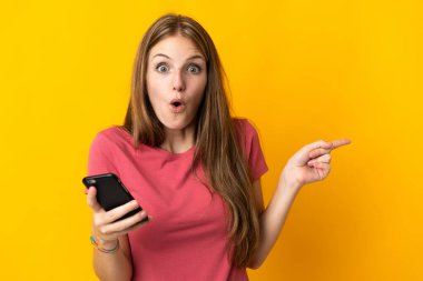 Sarı arka planda izole edilmiş cep telefonu kullanan genç kadın şaşırmış ve işaret ediyor.