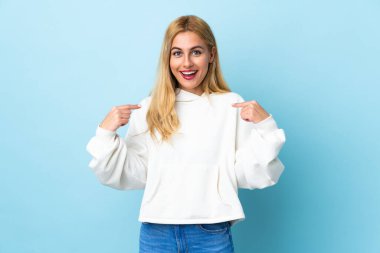 Genç Uruguaylı sarışın kadın, yalıtılmış mavi arka planda, şaşırtıcı bir yüz ifadesi var.