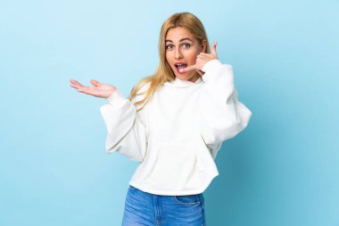 Genç Uruguaylı sarışın kadın izole edilmiş mavi arka planda telefon jestleri yapıyor ve kuşkulu