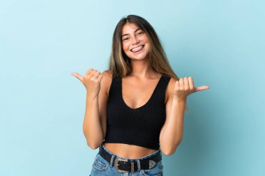 Mavi arka planda izole edilmiş genç beyaz kadın başparmakları havada ve gülümsüyor.