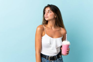 Mavi arka planda izole edilmiş çilekli milkshake 'li genç bir kadın yan tarafa bakıyor.