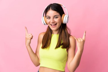 Pembe arka planda izole edilmiş genç beyaz kadın rock hareketleri yaparak müzik dinliyor.
