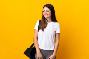 Sarı arka planda izole edilmiş spor çantalı genç bayan yana bakıyor ve gülümsüyor.