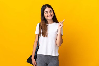 Spor çantalı genç bayan sarı arka planda izole edilmiş ve en iyinin işareti olarak parmağını kaldırıyor.