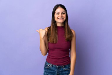 Mor arka planda izole edilmiş genç beyaz kadın bir ürünü sunmak için kenarı işaret ediyor.