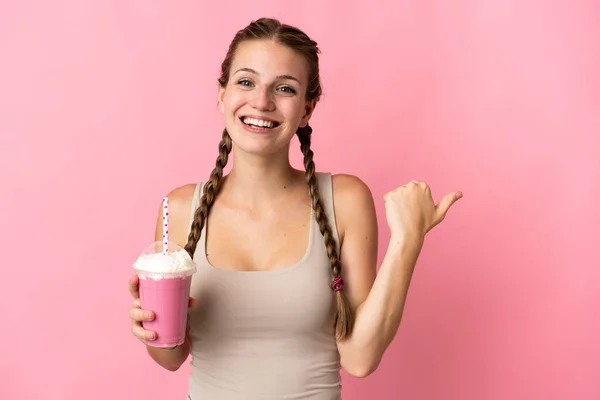 ピンクの背景に描かれたイチゴのミルクシェイクを持つ若い女性が商品を提示するために側面を指している — ストック写真