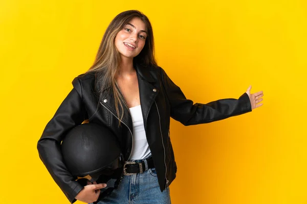 脇に手を伸ばし黄色の背景に隔離されたオートバイのヘルメットを持っている女性が来て招待するために — ストック写真