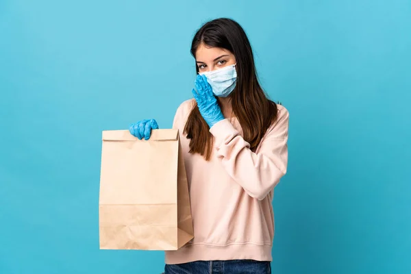 マスクでコロナウイルスから保護し 青い背景に隔離された食料品の買い物袋を持っている若い女性が何かをささやく — ストック写真