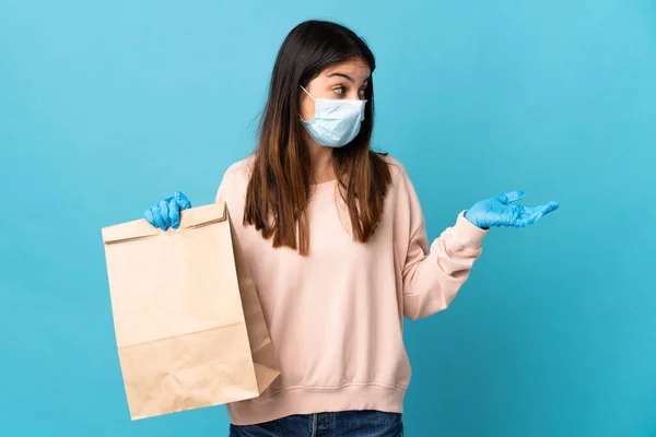 マスクでコロナウイルスから保護し 驚きの表情で青の背景に隔離された食料品店の買い物袋を保持若い女性 — ストック写真