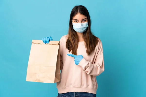 マスクでコロナウイルスから保護し 青の背景に隔離された食料品の買い物袋を保持し それを指す若い女性 — ストック写真