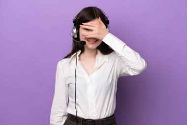 Telemarketer Russian woman working with a headset isolated on purple background covering eyes by hands and smiling