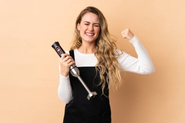 Young brazilian woman using hand blender isolated on beige background doing strong gesture