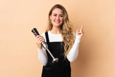 Bej arka planda izole edilmiş el blenderi kullanan genç Brezilyalı kadın harika bir fikre işaret ediyor.