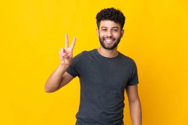 Sarı arka planda izole edilmiş genç Faslı adam gülümsüyor ve zafer işareti gösteriyor