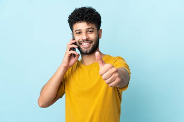 Young Moroccan man isolated on blue background keeping a conversation with the mobile while doing thumbs up