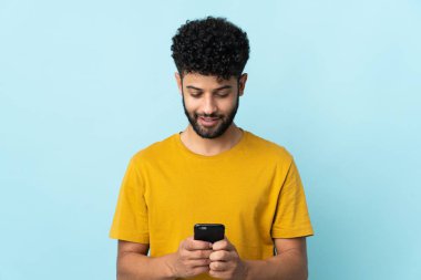 Genç Faslı bir adam mavi arka planda izole edilmiş. Cep telefonuyla mesaj ya da e-posta gönderiyor.