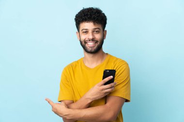 Young Moroccan man isolated on blue background using mobile phone and pointing back