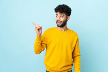 Faslı genç bir adam mavi arka planda izole edilmiş bir şekilde parmağını kaldırarak çözümü bulmaya çalışıyor.