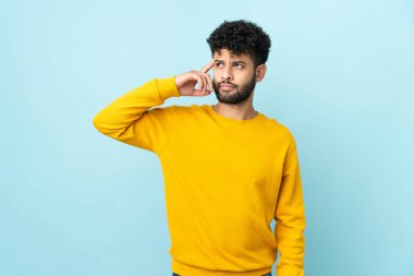 Faslı genç bir adam mavi arka planda izole edilmiş. Delilik jesti yapıyor. Parmağını kafaya koyuyor.