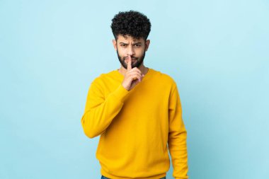 Faslı genç bir adam mavi arka planda izole edilmiş. Parmağını ağzına koyduğunda sessizlik işareti gösteriyor.