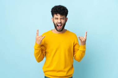 Faslı genç bir adam mavi arka planda izole edilmiş boynuz hareketi yapıyor.