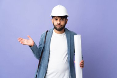 Young architect Moroccan man with helmet and holding blueprints over isolated background having doubts while raising hands