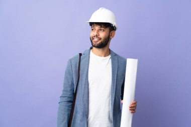 Young architect Moroccan man with helmet and holding blueprints over isolated background thinking an idea while looking up